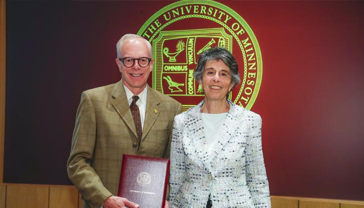 Chair Mayeron and Interim Chancellor McMillan posing for a photo with the Board of Regents seal on a maroon background behind them.